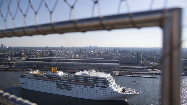 Cruise ship Costa neoRomantica, operated by Carnival Corp., sails on the IJ river as Amsterdam central railway station stands beyond in Amsterdam, Netherlands, on Thursday, Aug. 25, 2016. The district of Amsterdam Noord is taking off as soaring housing costs across the city and the promise of a new subway line tempt residents to brave the bottlenecked journey across the IJ river in search of better values or lower rents.